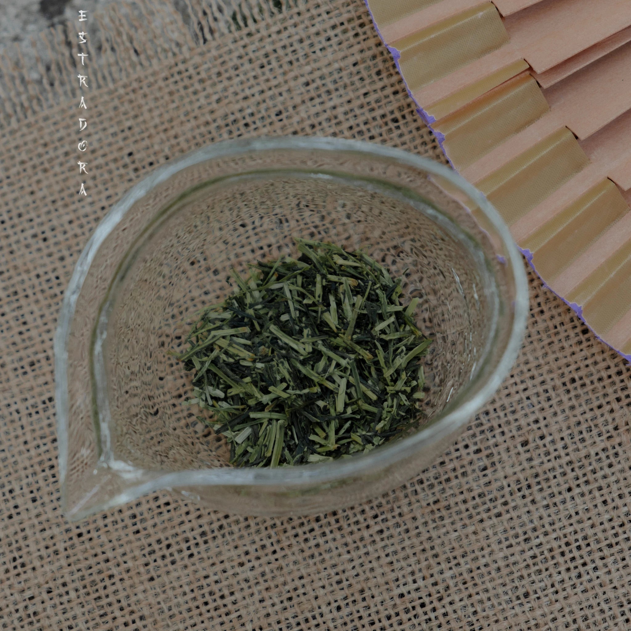 Loose leaf Japanese Kukicha green tea in glass teapot, close-up on natural fabric background - Estradora