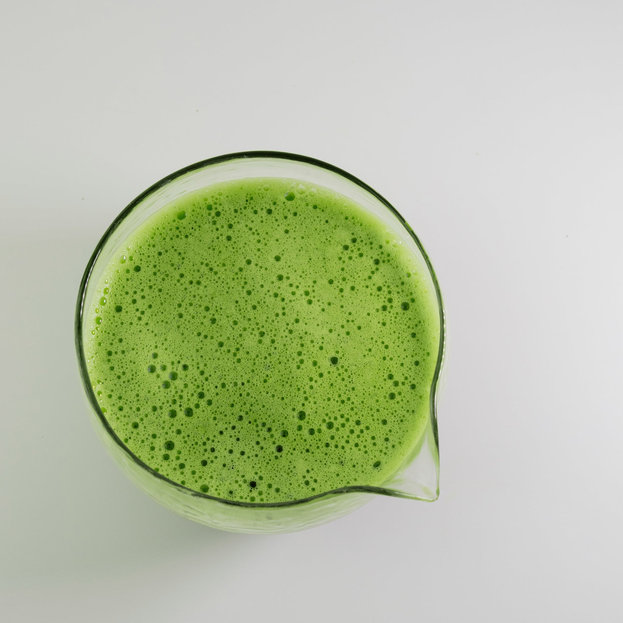 Top-down view of whisked Kasumi ceremonial matcha in a glass pitcher, smooth green foam on a white surface.
