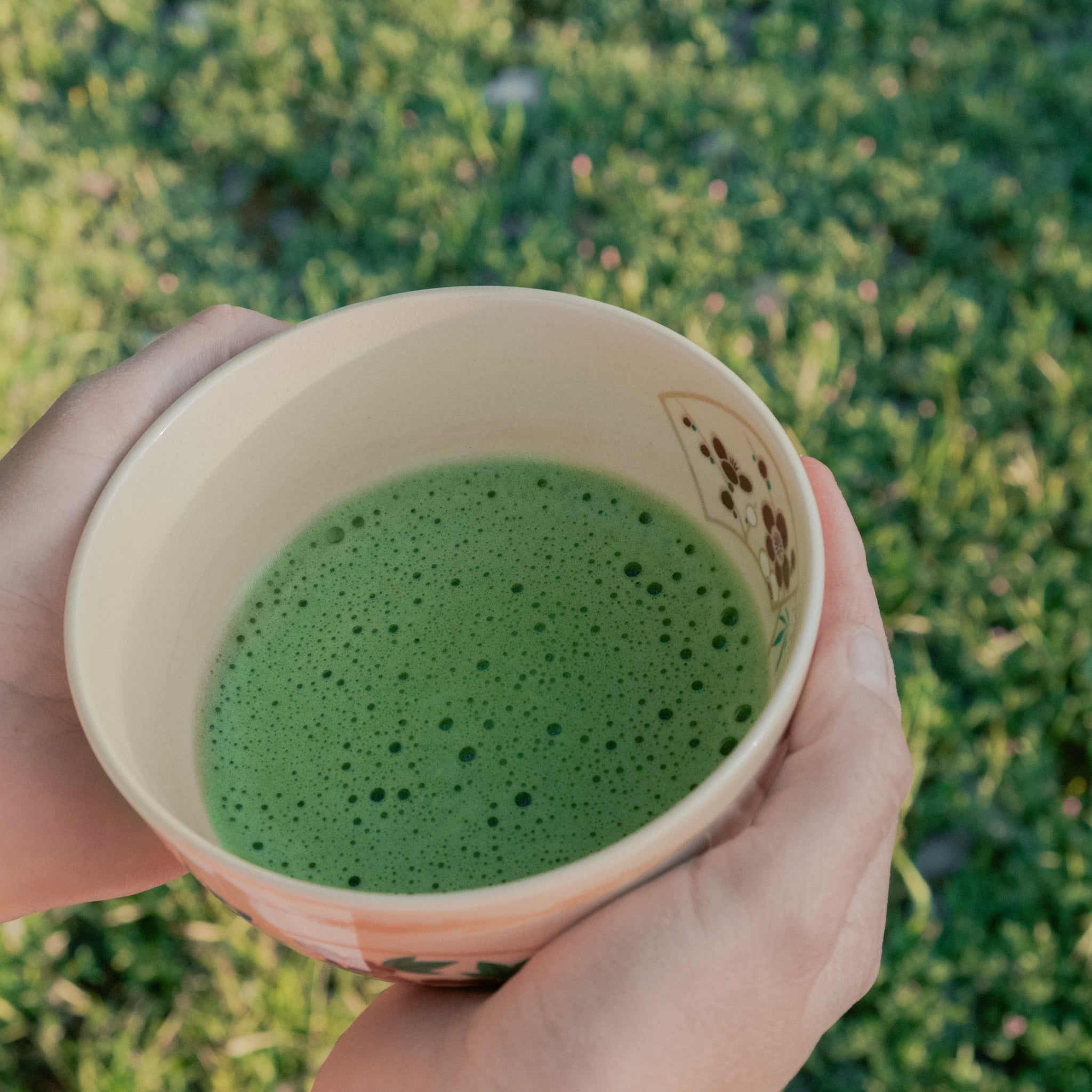 Hand holding a Japanese chawan with freshly whisked Kasumi ceremonial matcha, fine green foam, garden background.