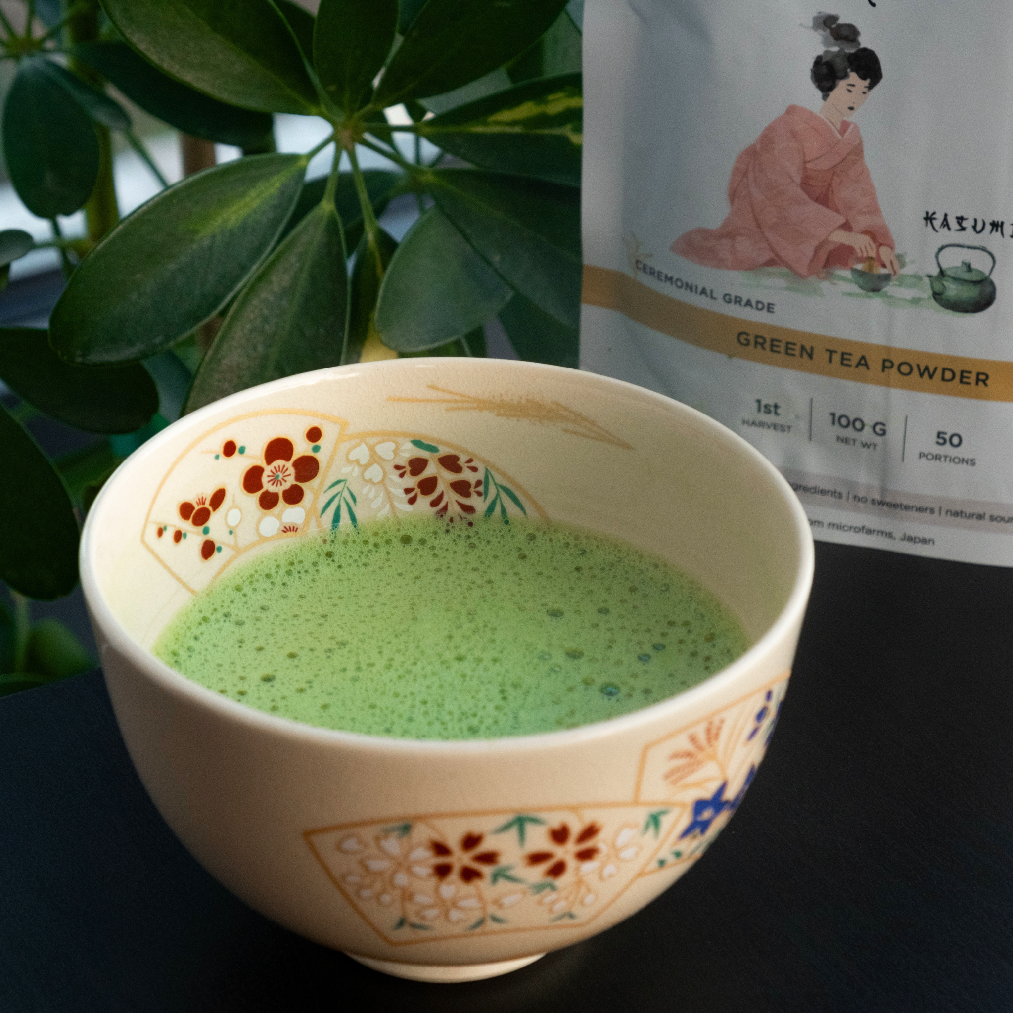 KASUMI matcha in a hand-painted ceramic bowl, its packaging and a potted plant in the background.